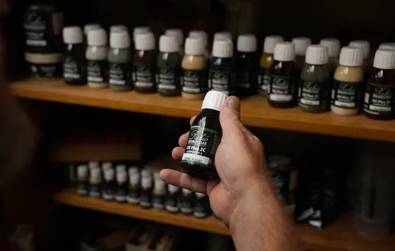 A hand is holding a small bottle of Oil Plus 2C in front of a cabinet filled with rows of Oil Plus 2C bottles in different colours