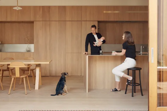 A man holding a baby is standing at a kitchen counter and looking at his dog. A woman is seated on a bar stool. The entire dining and kitchen area are wooden surfaces in a light, natural tone and are protected by the food and toy safe wood oils from Rubio Monocoat.